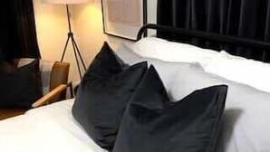 Uma cama bem arrumada com almofadas escuras e claras, uma luminária e um quadro na parede.