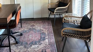 Una habitación con una mesa de comedor de madera, dos sillas de mimbre, un tapete estampado y un amplio ventanal con persianas.