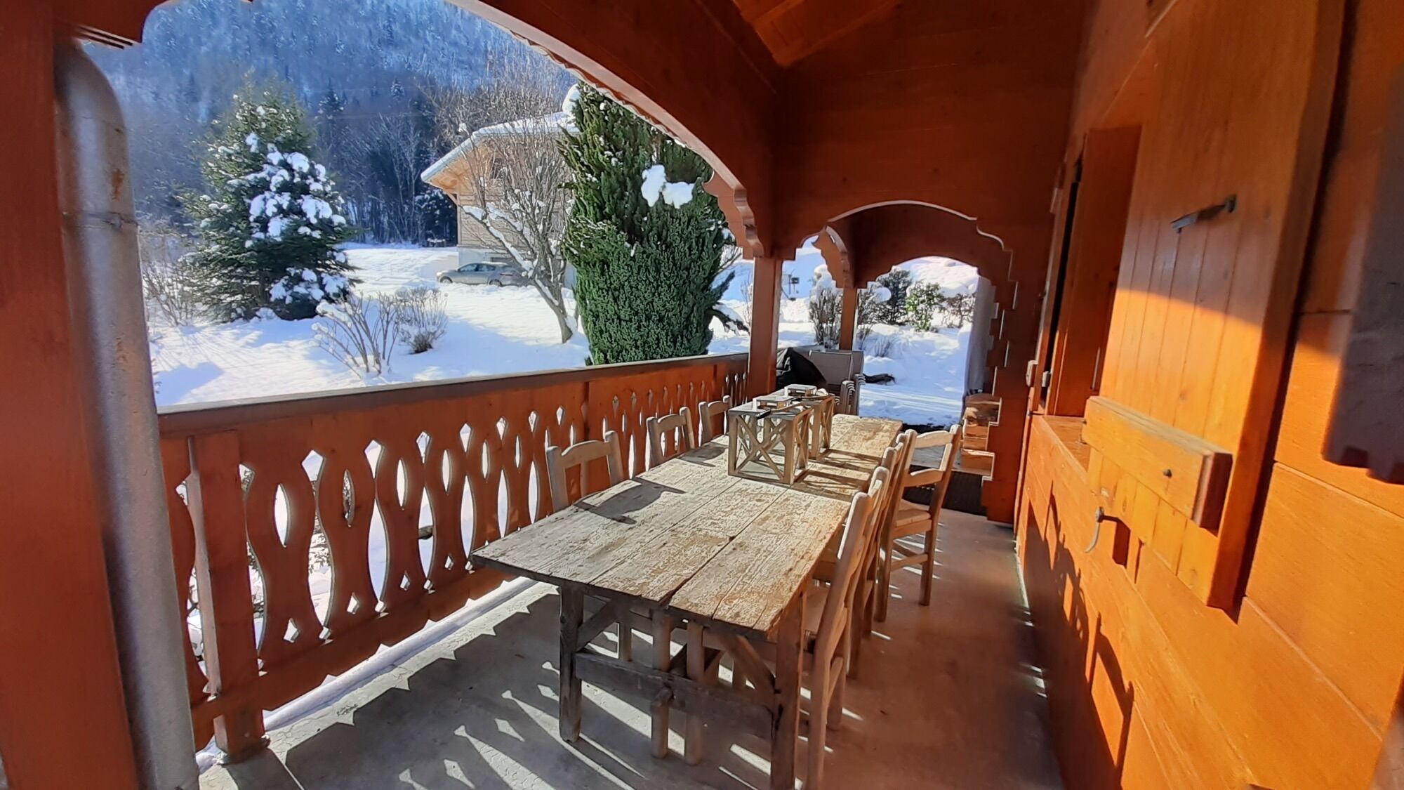 A wooden deck with a table and chairs, overlooking a snowy landscape with trees and a building.