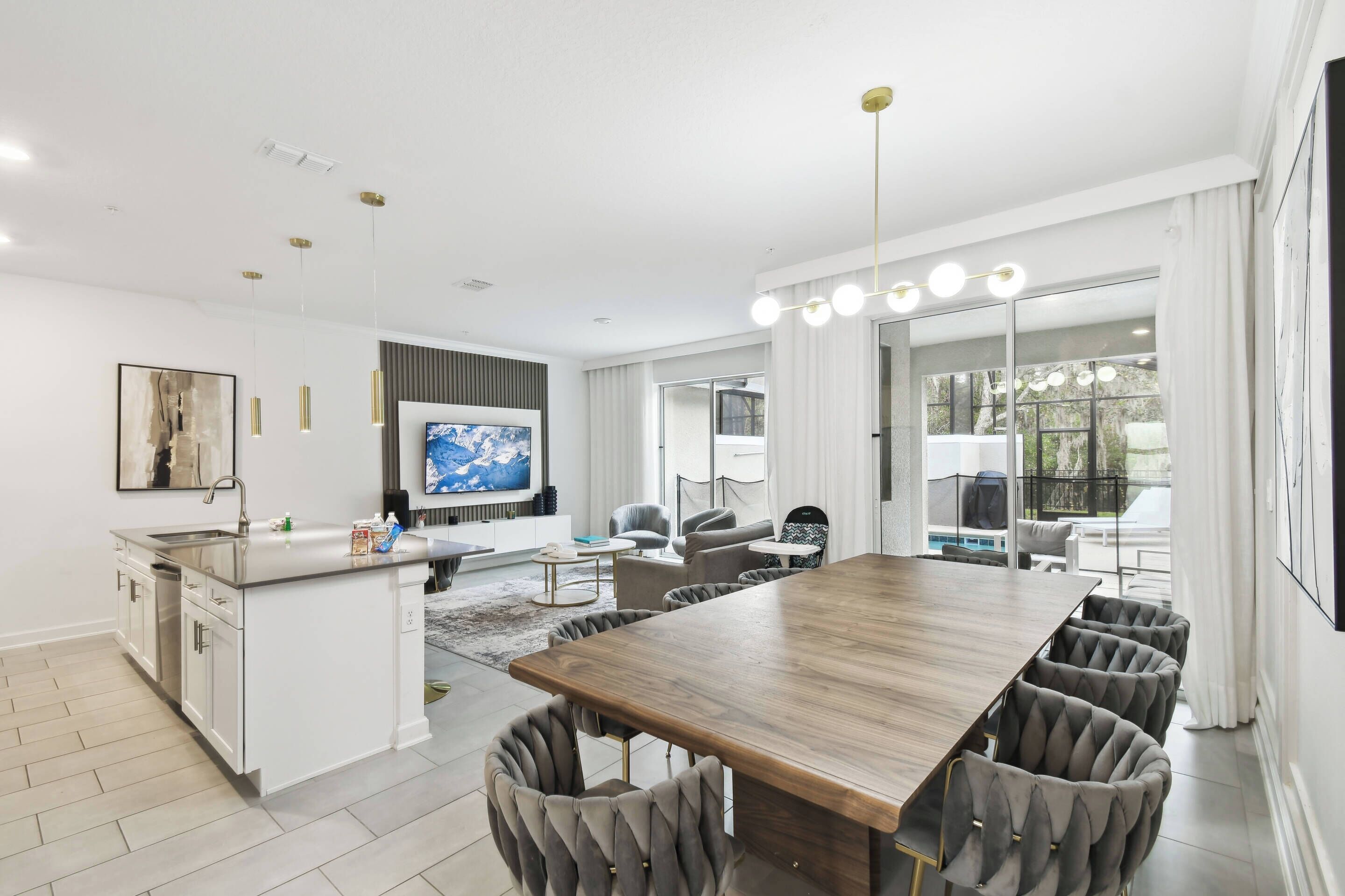 Una cocina moderna con una isla central, un comedor con mesa de madera y sillas tapizadas, y una sala de estar con sofá y televisor.