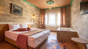 A bedroom with a wooden headboard, a bed with pink bedding, a bedside table, a large window with curtains, and a built-in bathtub. - Arton In Cappadocia (Avanos)