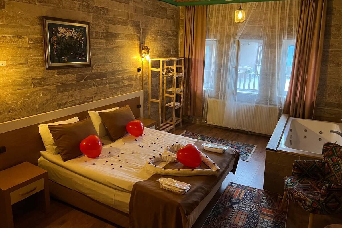 A bedroom with a bed, a nightstand, a bedside table, a window with curtains, a wall-mounted shelf, and a bathroom with a bathtub.