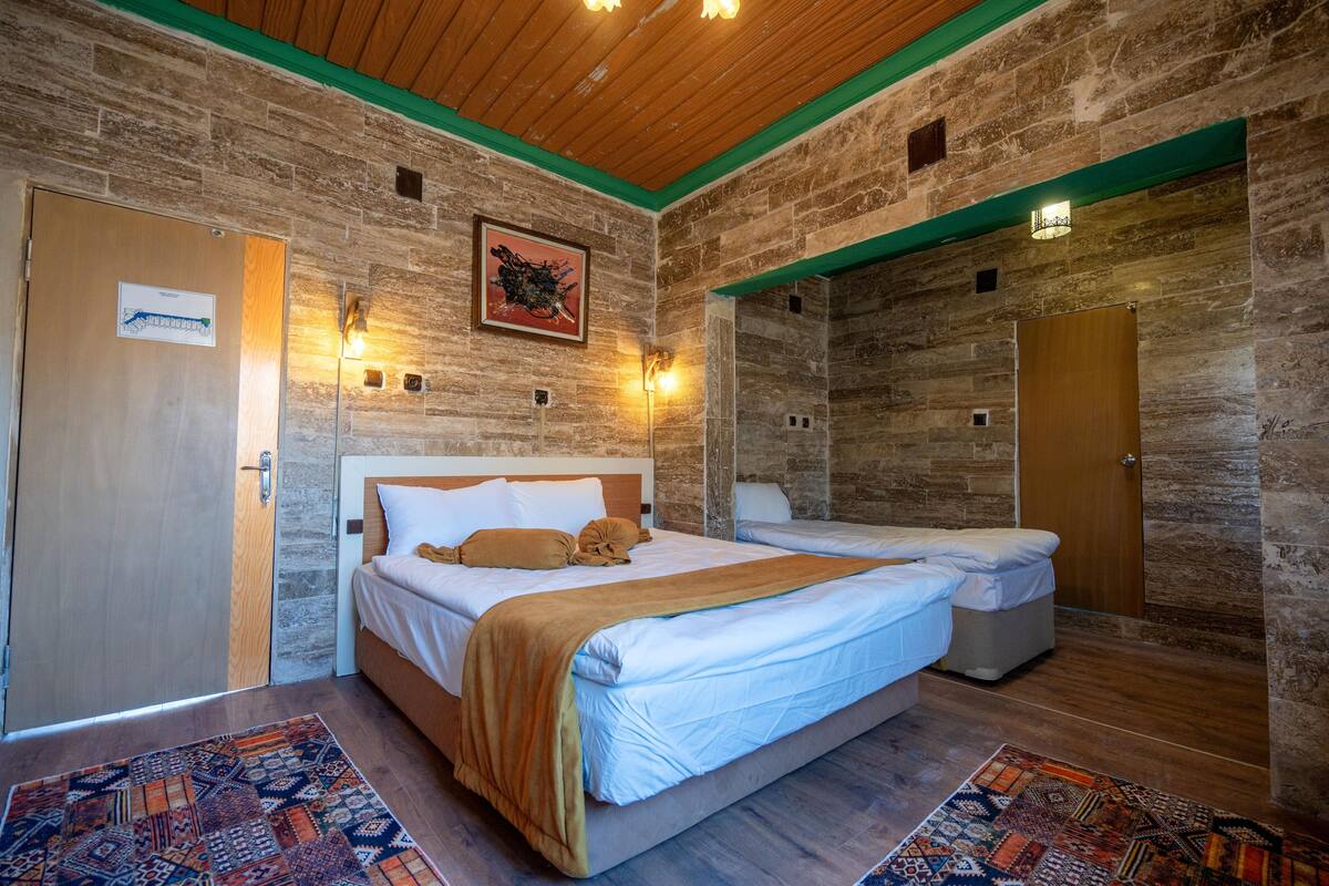 A hotel room with a large bed, a smaller bed, a wooden ceiling, stone walls, and a patterned rug.