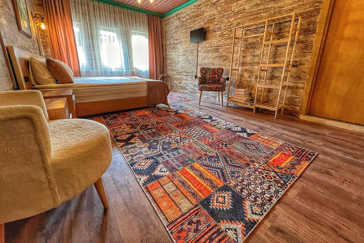 A cozy room with a bed, a chair, a patterned rug, and a wooden ladder against a stone wall.