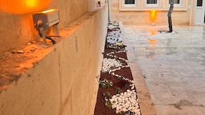 A courtyard with a pathway lined with pebbles and flowers, a wall with a light fixture, and a building with decorative windows and a balcony. - Arton In Cappadocia (Avanos)