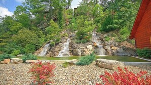 A natural landscape with a waterfall, surrounded by trees and a wooden cabin. - Cozy Bear Heaven: Cuddle Up in the Smokies (Sevierville)