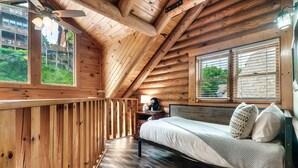A wooden cabin bedroom with a bed, a nightstand, a lamp, and a window overlooking greenery.