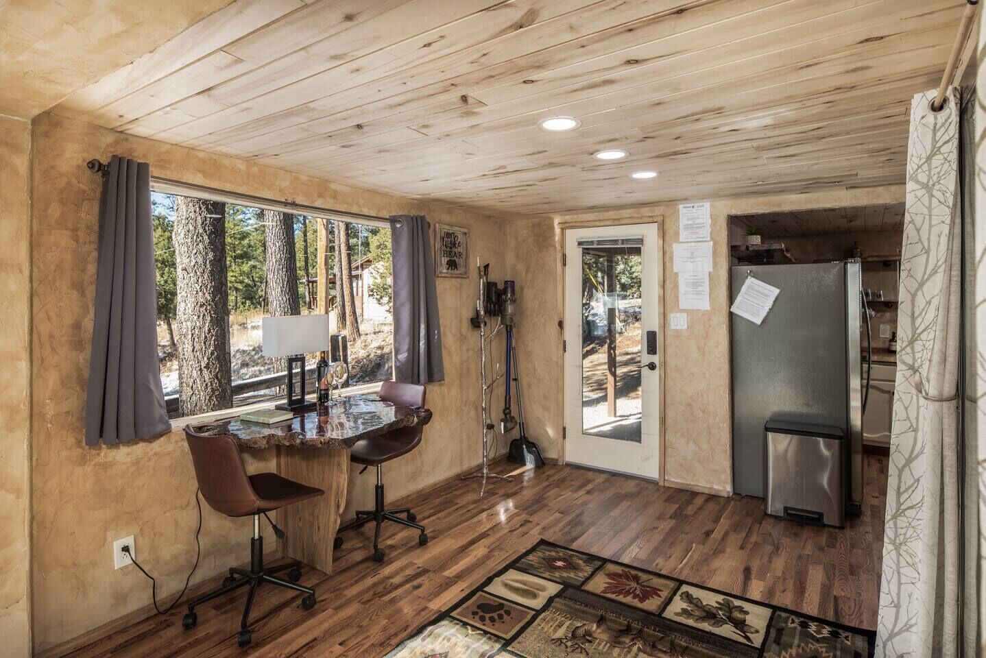 A rustic cabin interior with a wooden ceiling, a dining area with a table and chairs, a refrigerator, and a door leading outside.