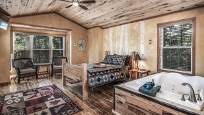 A cozy bedroom with a wooden ceiling, a large bed with a patterned quilt, a bedside table with a lamp, and a window with blinds. - Redwood in the Historic Upper Canyon (Ruidoso)