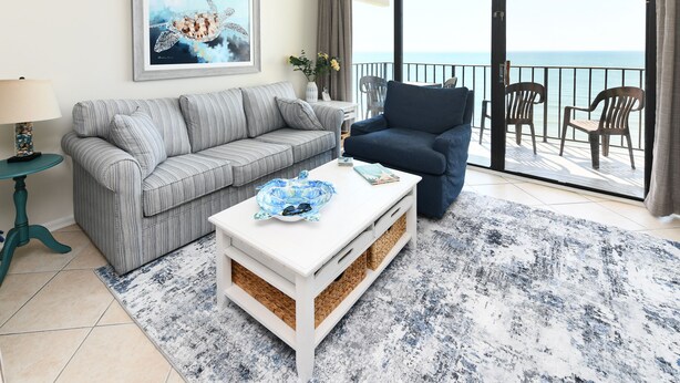 Ein Wohnzimmer mit einem grauen Sofa mit Streifen, einem blauen Sessel, einem weißen Couchtisch mit einer blauen Schale und einem großen Fenster mit Blick auf das Meer.