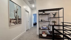 A hallway with a framed picture of a cactus, a black metal shelf unit with books and a plant, and a white door.