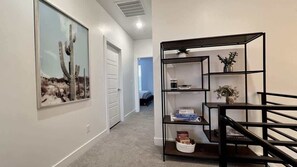 A hallway with a framed picture of a cactus, a black metal shelf unit with books and a plant, and a white door. - The Lofts #44 | Townhome Pool Resort (Saint George)