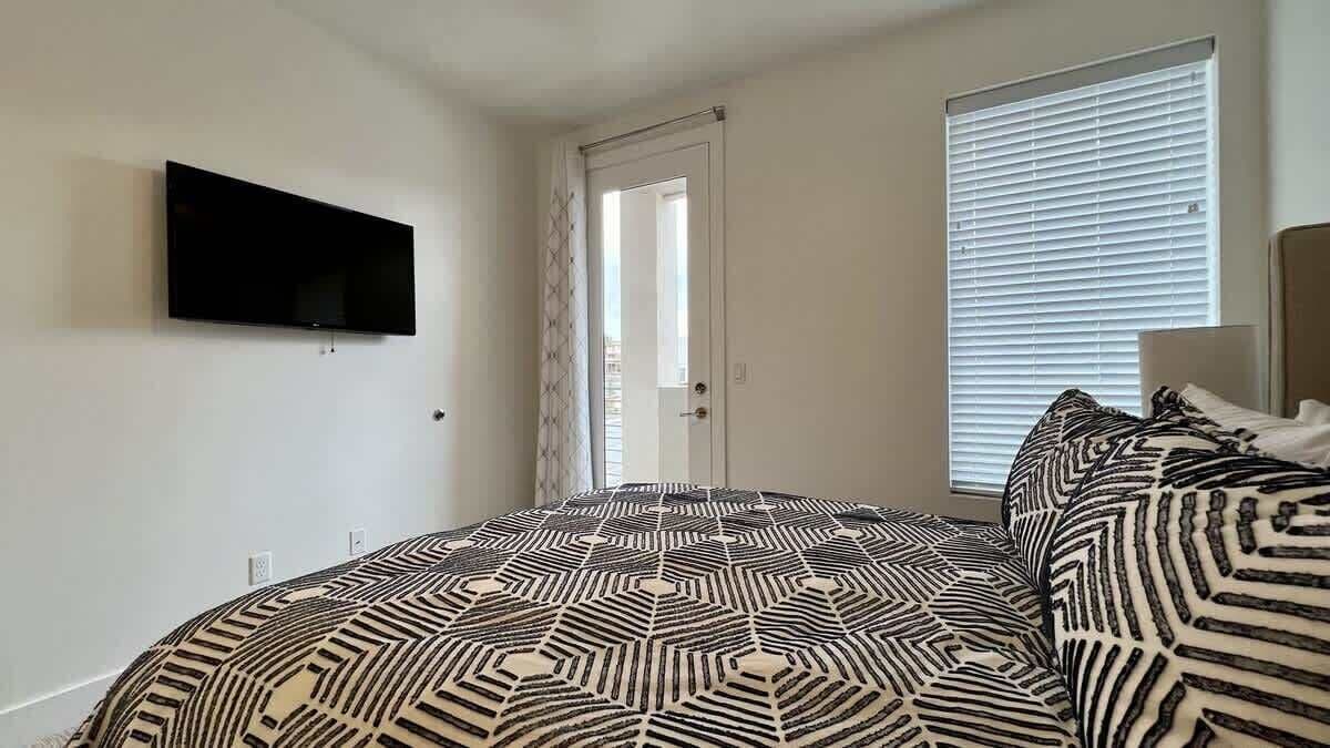 A bedroom with a bed, a television mounted on the wall, a door, and a window with blinds.
