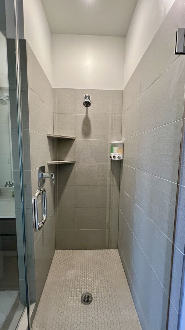 A walk-in shower with a glass door, a handheld showerhead, a soap dispenser, and a small shelf.
