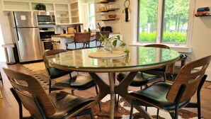A modern kitchen with a glass dining table surrounded by leather chairs, a cowhide rug, and a view of greenery through a large window. - Waterfront Cottage with a sand beach! (Green Bay)