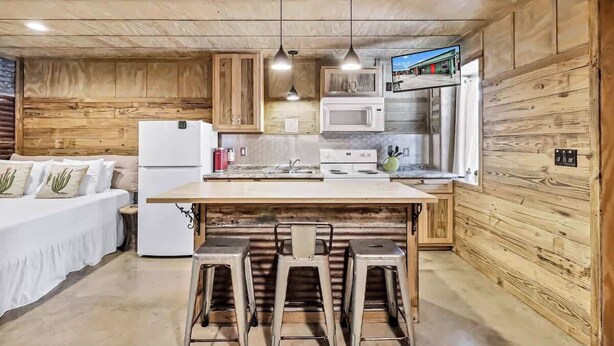 A compact kitchen with wooden walls, a white refrigerator, a microwave, and a sink. There are bar stools and a small table in the center. - Casas Coastal Experience Cabin A (Rockport)