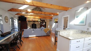 A spacious living area with a stone fireplace, wooden beams, a dining table with chairs, and a kitchen with white cabinets and granite countertops. - Updated 3 Bedroom Offering Hot Tub and Large Yard (2360) (Brewster)