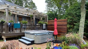 A hot tub on a wooden deck with a pergola overhead, surrounded by a garden with various plants and flowers. - Updated 3 Bedroom Offering Hot Tub and Large Yard (2360) (Brewster)