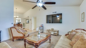 A spacious living room with a ceiling fan, a large flat-screen TV, a glass coffee table, and a sofa with patterned cushions. - 8 Mi to State Farm Stadium! Poolside Retreat (Peoria)