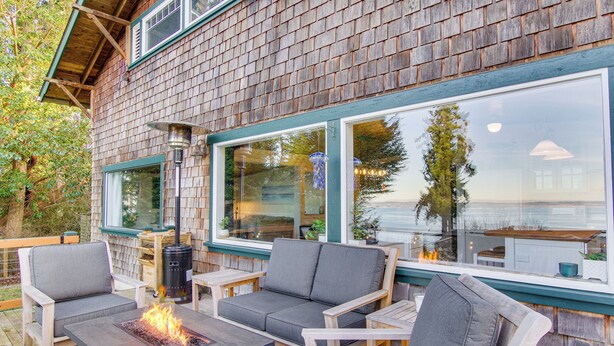 An outdoor deck with a fire feature, seating, and large windows overlooking a body of water. - Puget Sound Cottage w/ Private Community Beach! (Coupeville)