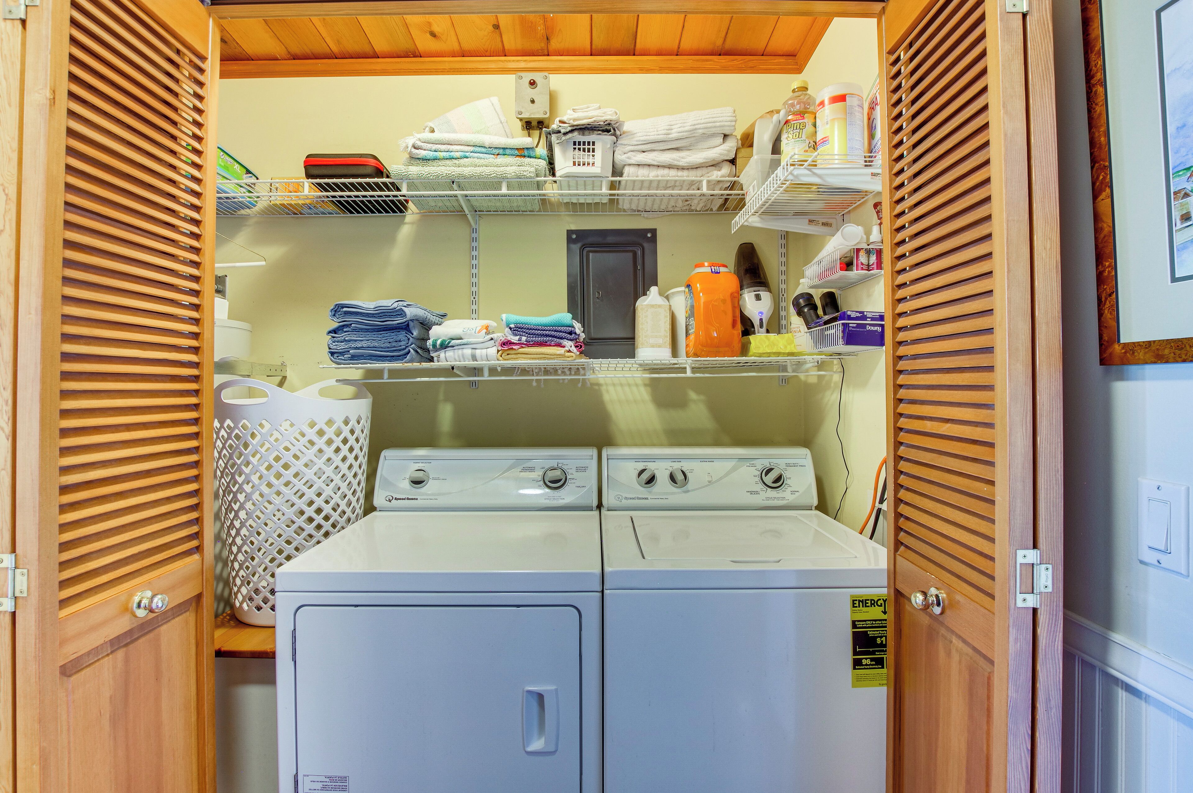 A laundry room with two stacked washers and dryers, shelves with folded towels and laundry supplies, and a wooden door with a louvered design.