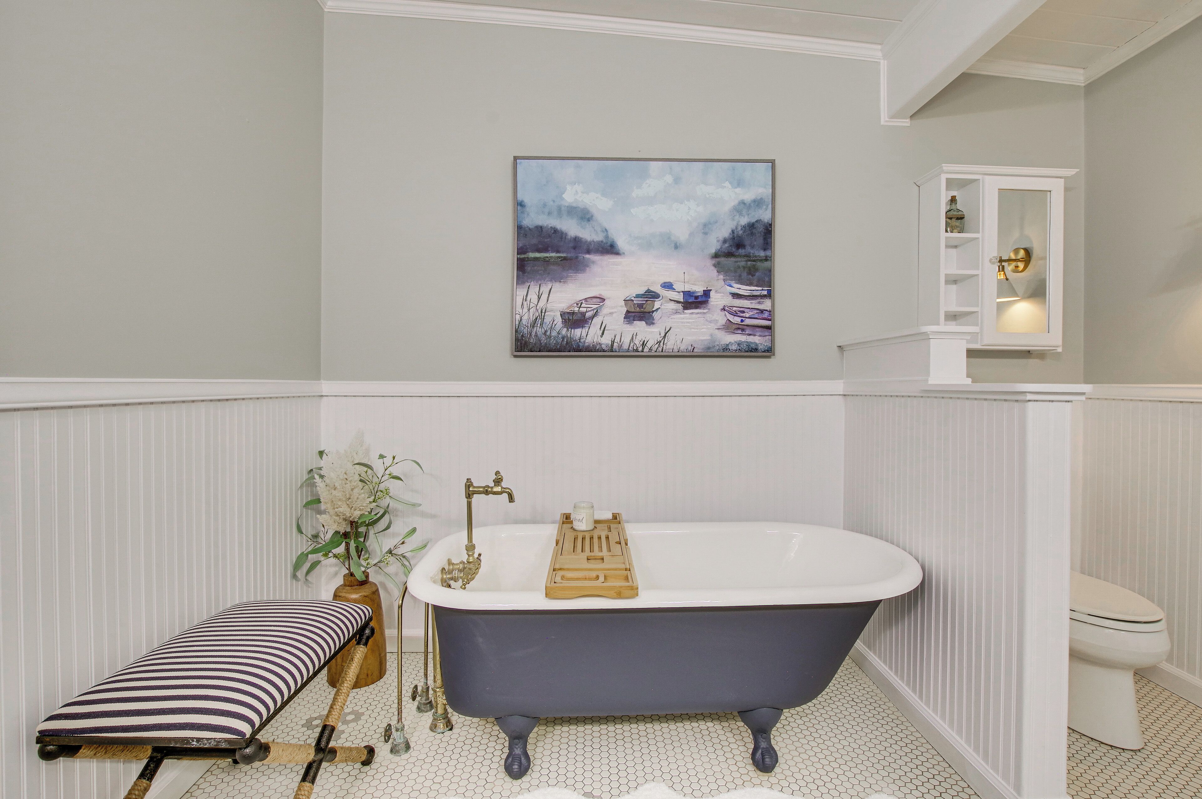 A bathroom with a freestanding bathtub, a wooden bench, a vase with flowers, and a painting of boats on the wall.