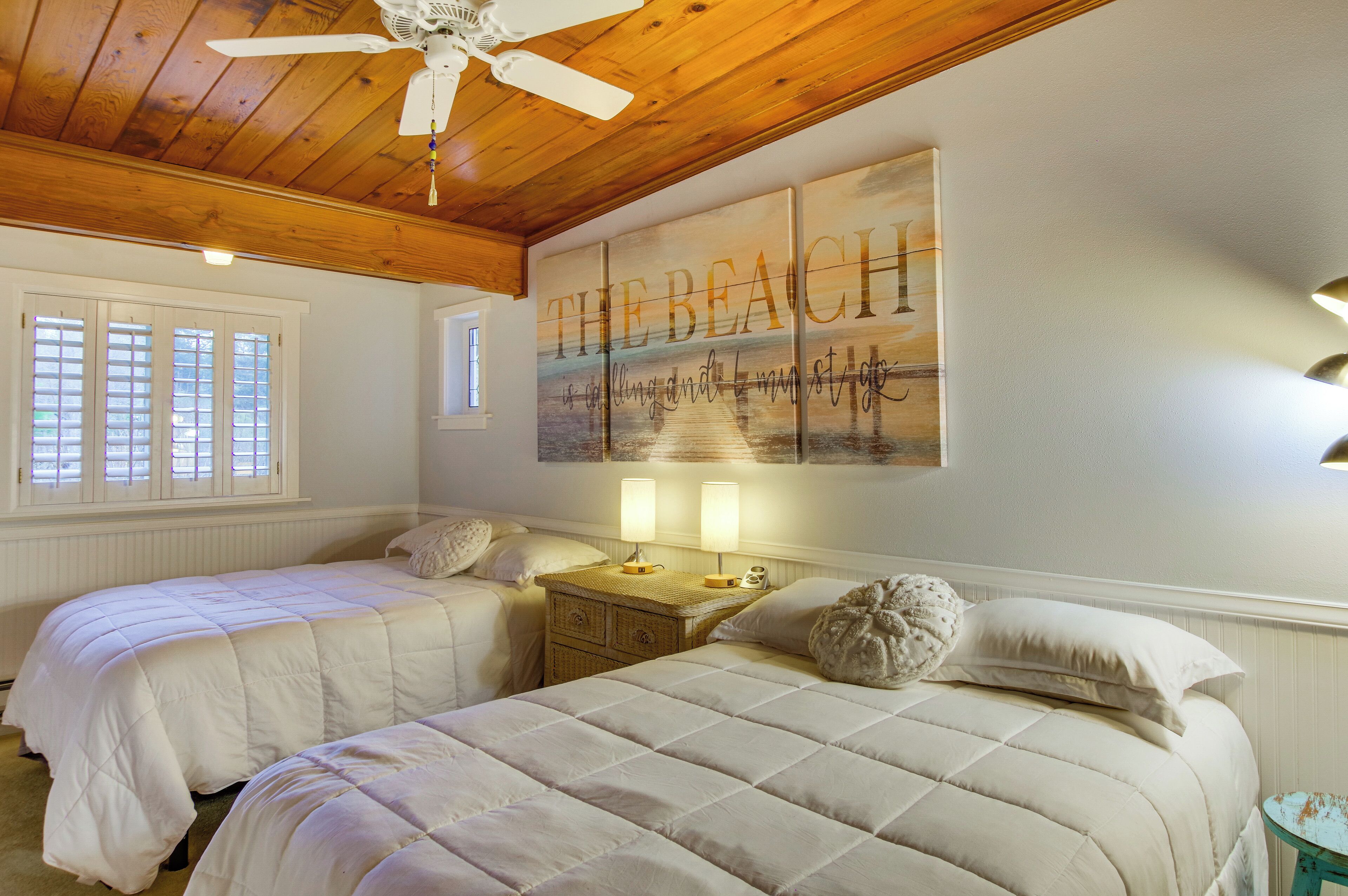 A bedroom with two beds, a ceiling fan, a beach-themed wall art, and a window with blinds.