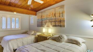 A bedroom with two beds, a ceiling fan, a beach-themed wall art, and a window with blinds.