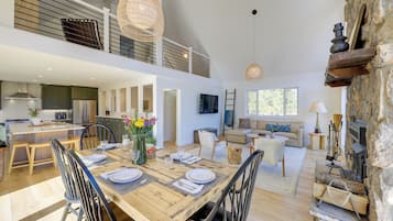 A spacious open-plan living area with a wooden dining table set for four, a stone fireplace, and a kitchen in the background.