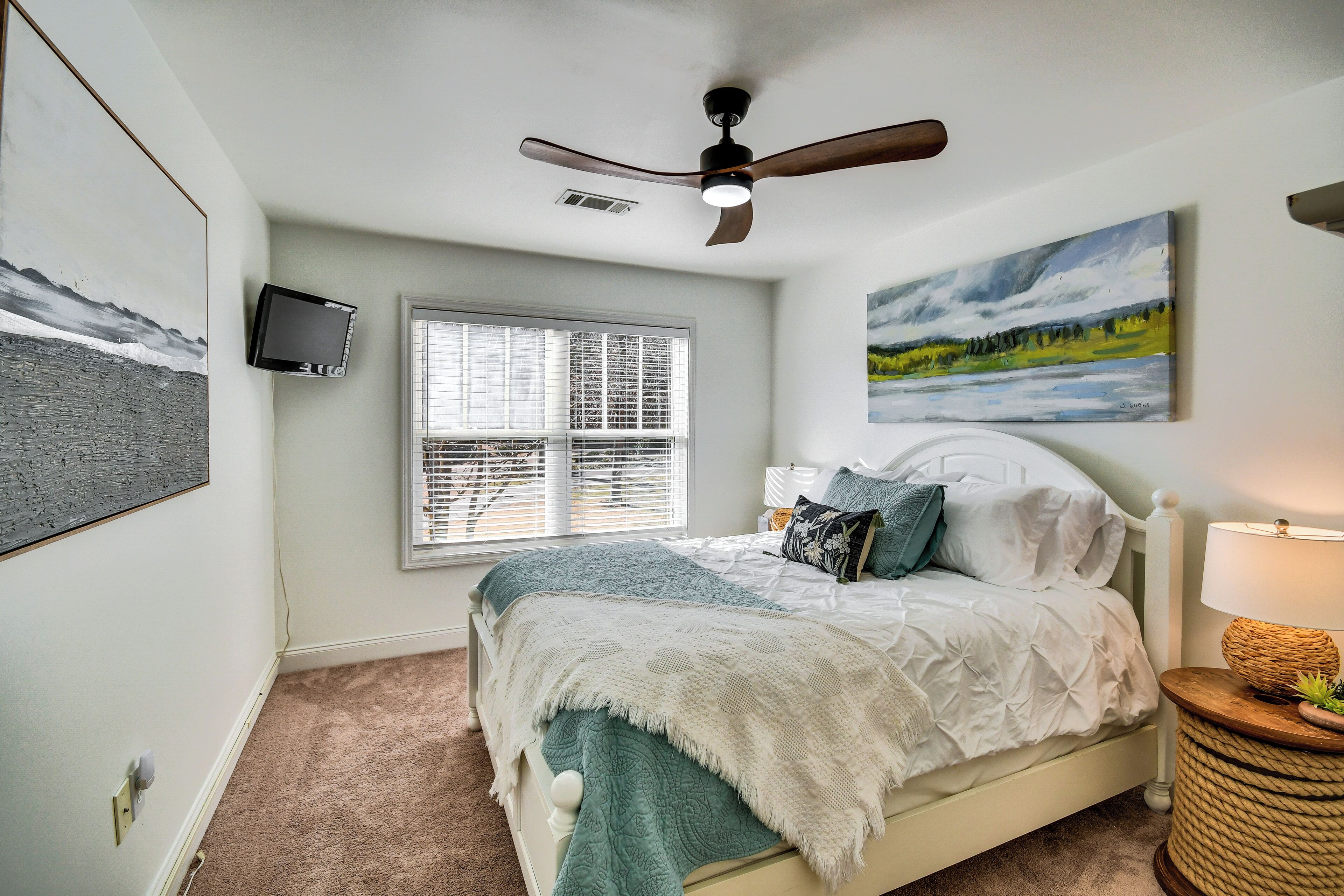 A bedroom with a bed, a ceiling fan, a window, a painting, and a lamp.
