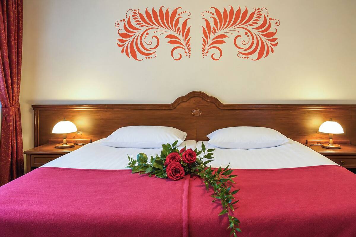 Habitación de hotel con cabecera de madera, dos lámparas de noche, cama con ropa de cama blanca y roja, y un arreglo floral sobre la cama.