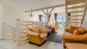 Un salón amplio con dos sofás y una mesa de centro, una escalera con baranda de madera, y grandes ventanas que dan a zonas verdes.