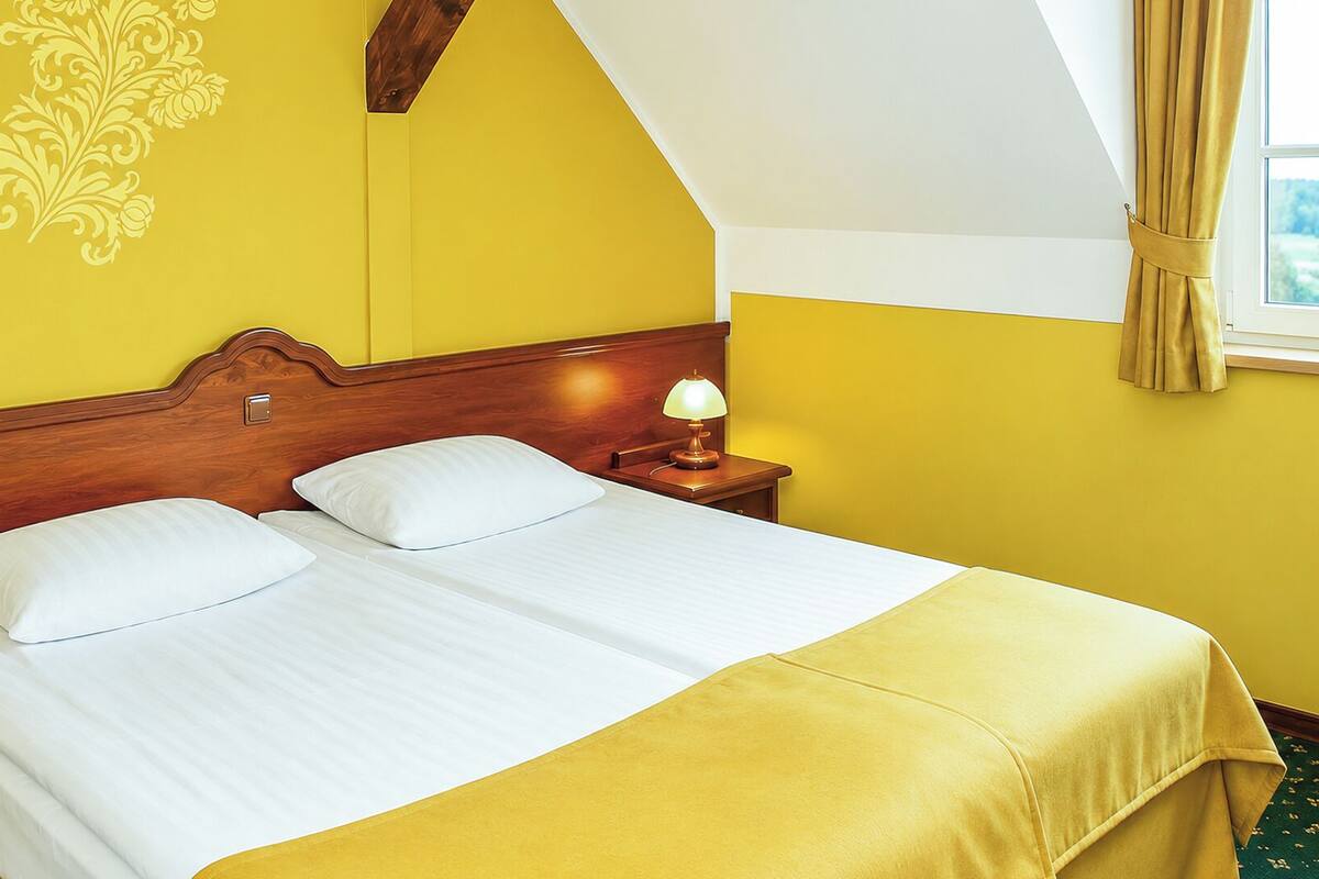 Una habitación de hotel con cabecera de madera, dos camas blancas, una lámpara de noche y una ventana con cortinas.