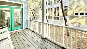 Um deck de madeira com guarda-corrimão branco, uma porta verde e vista para uma casa com várias janelas e uma árvore.
