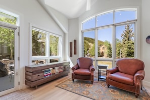 A spacious room with large windows, a leather armchair, a wooden console table, and a view of a forested area. - Legends 10 4br 4ba 4 Bedroom Townhouse by RedAwning (Copper Mountain)