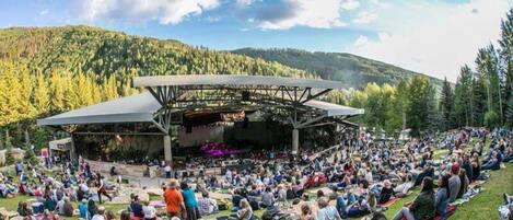 An outdoor amphitheater with a stage, surrounded by a grassy area with people seated.
