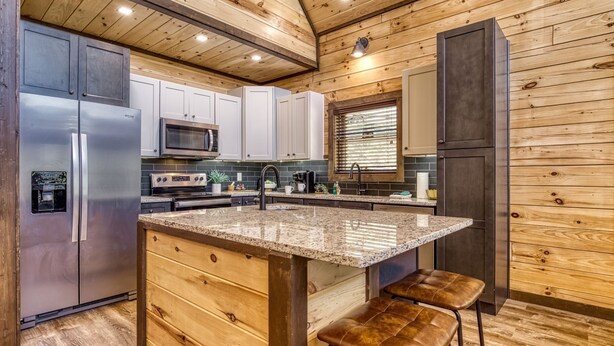 A modern kitchen with wooden paneling, stainless steel appliances, and a granite island with bar stools. - Er29 - Pop's Perch 2 Bedroom Cabin by RedAwning (Pigeon Forge)