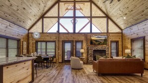 A spacious interior with a high ceiling, wooden walls, a stone fireplace, and a dining area with a view of the outdoors. - Er29 - Pop's Perch 2 Bedroom Cabin by RedAwning (Pigeon Forge)