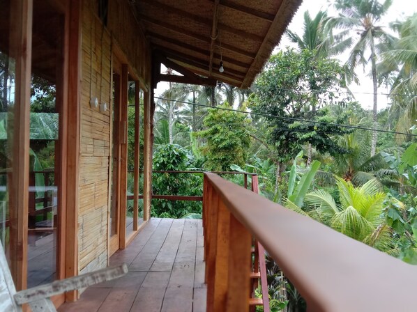 A wooden balcony with a thatched roof, overlooking a lush garden with various tropical plants and palm trees.