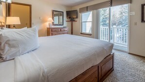 A bedroom with a large bed, a dresser, a television, and a balcony with a view of snow-covered trees.