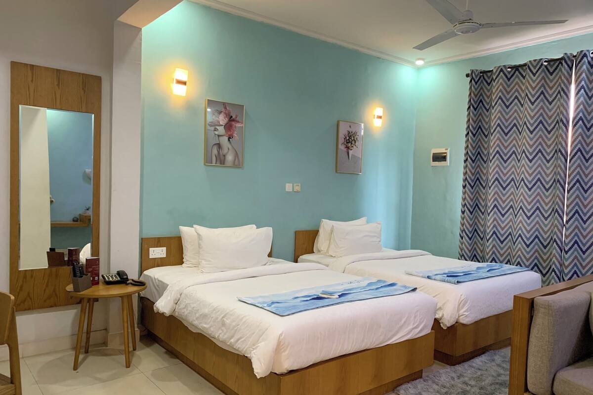 A hotel room with two beds, a desk, and a chair. There are wall-mounted light fixtures and a ceiling fan.