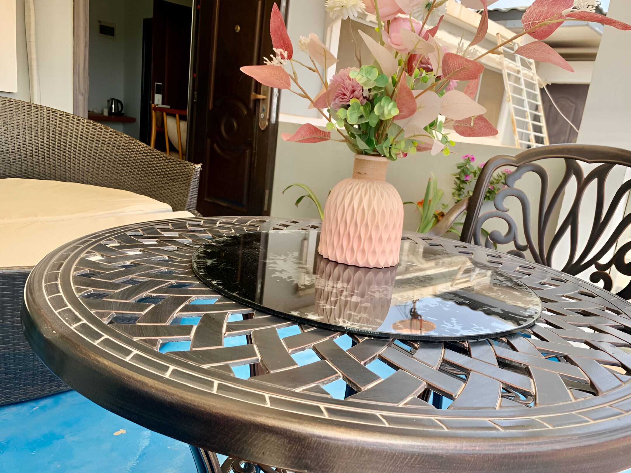 A round outdoor table with a glass top and a woven metal frame, accompanied by a wicker chair and a cushion.