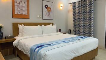 Un dormitorio de hotel con cabecera de madera, una cama grande con ropa de cama blanca y azul, una mesita de noche con un jarrón de flores y un aire acondicionado instalado en la pared.