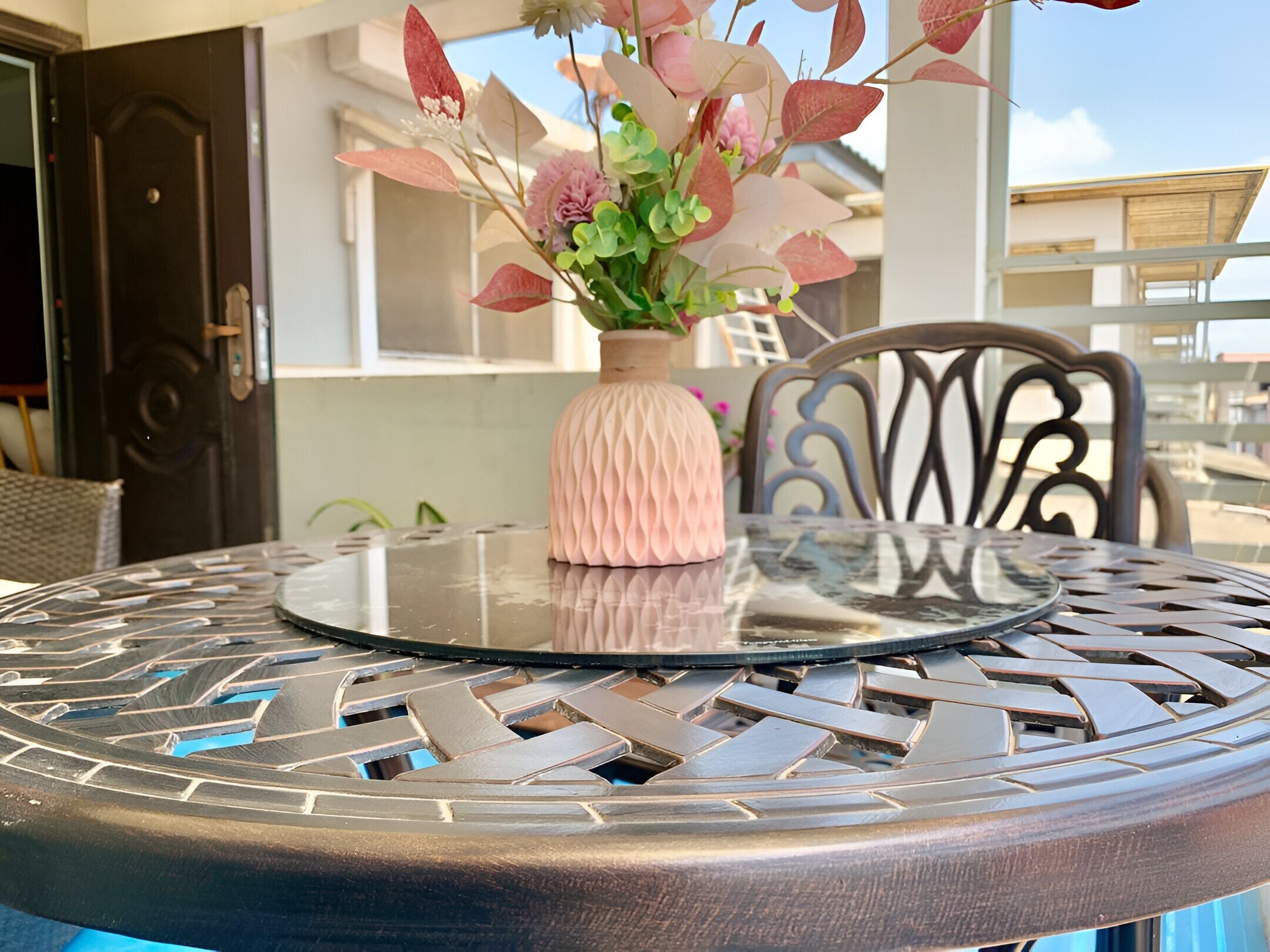 A round metal table with a glass top, a pink vase with flowers, and a wicker chair.