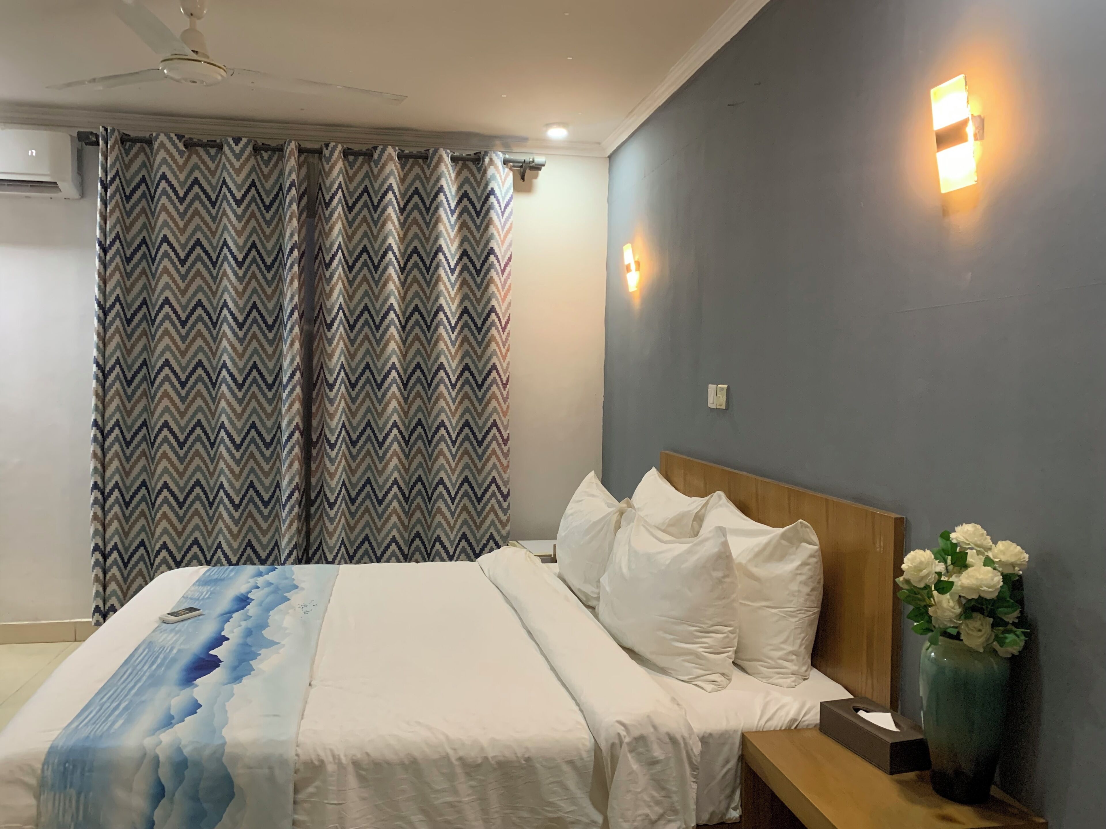 Een slaapkamer met een bed, kussens, een nachtkastje, een vaas met bloemen en een plafondventilator.
