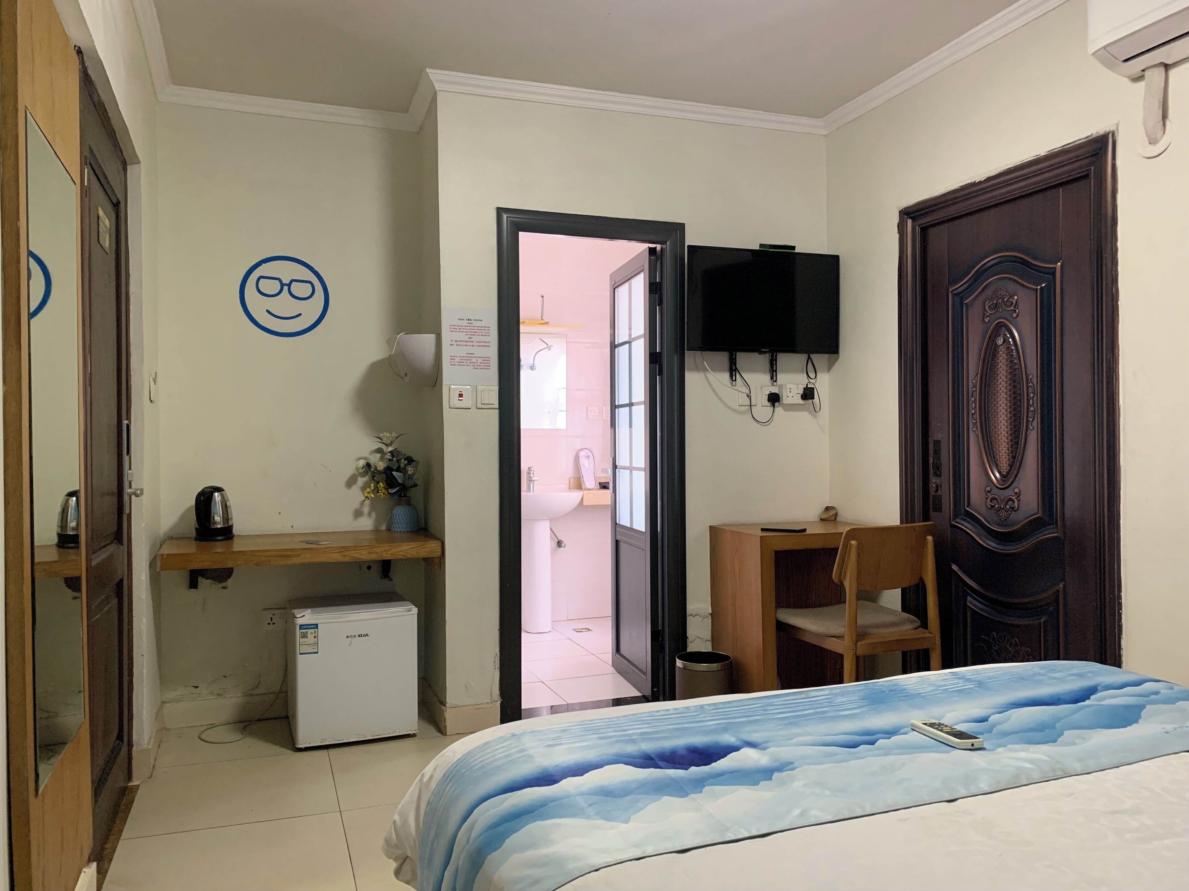 Una habitación de hotel con cama, escritorio, silla, refrigerador pequeño, televisor montado en la pared y puerta que da al baño.
