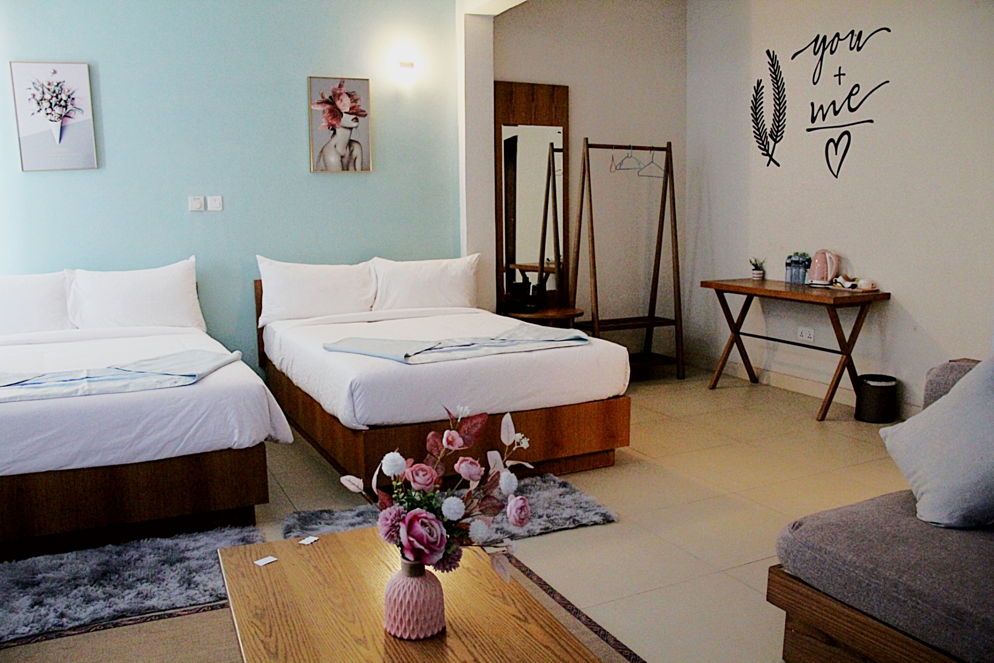 A hotel room with two beds, a wooden coffee table with a vase of flowers, a small table with a plant, and a wall decoration saying "you + me".