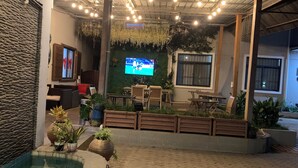 A covered outdoor seating area with a pool, potted plants, and a television screen. - Canwin Hotel (Accra)