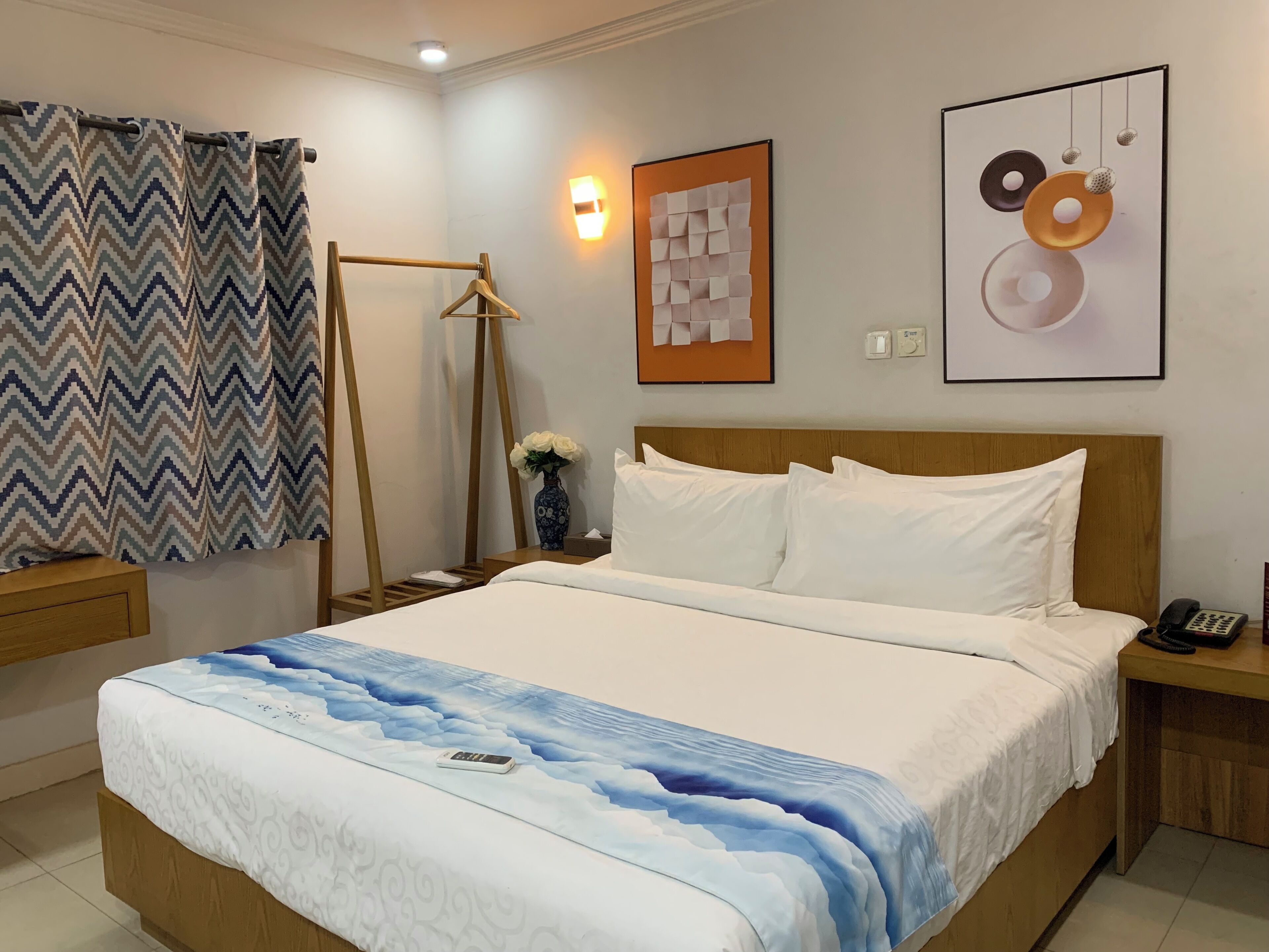 A neatly made bed with a blue and white patterned bedspread, a wooden headboard, and white pillows. There is a bedside table with a vase of flowers and a phone. The room has a chevron-patterned curtain, a wall-mounted light, and a framed abstract art piece.