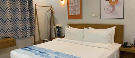 A neatly made bed with a blue and white patterned bedspread, a wooden headboard, and white pillows. There is a bedside table with a vase of flowers and a phone. The room has a chevron-patterned curtain, a wall-mounted light, and a framed abstract art piece.